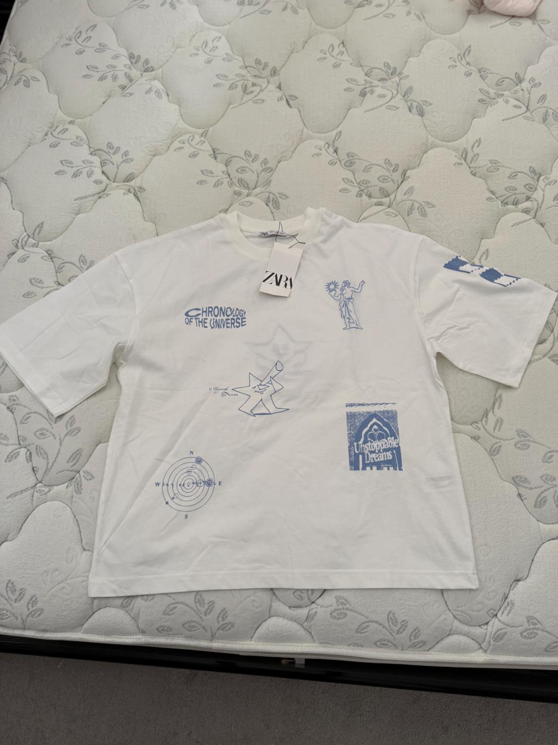 Front view of a white t-shirt with blue prints and the texts 'Chronicles of the Universe' and 'Unstoppable Dreams'.