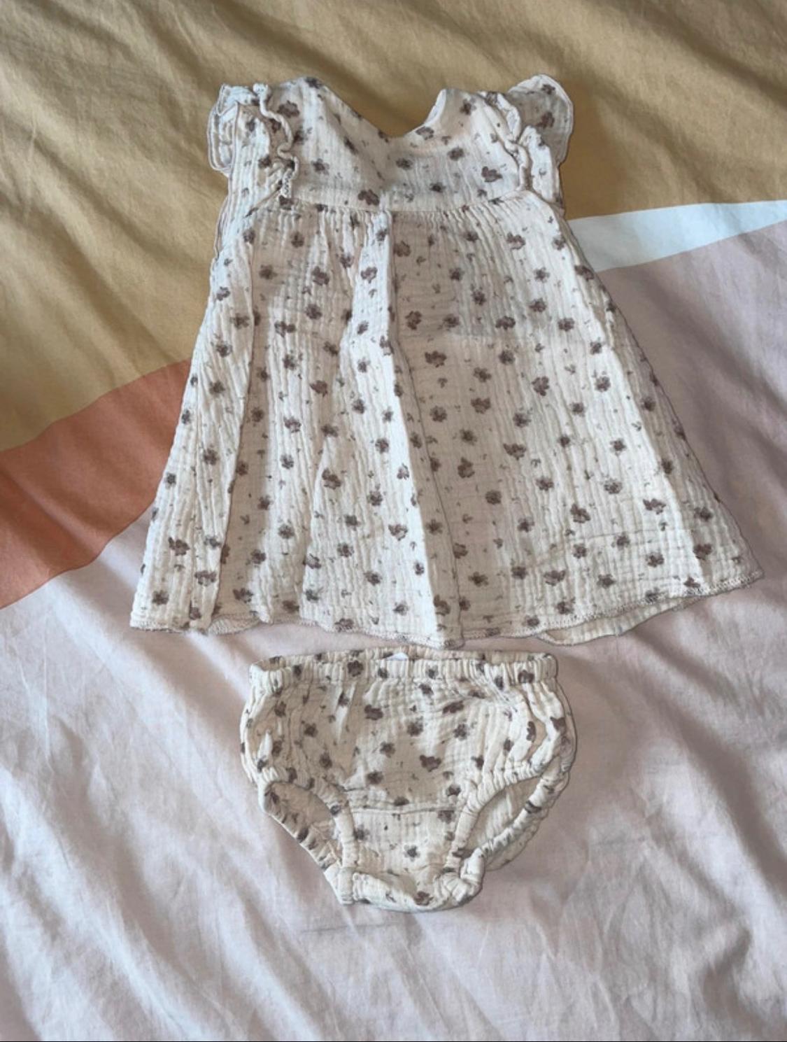 Baby dress and knickers with a floral print in beige and brown tones on a bed with soft-coloured sheets.