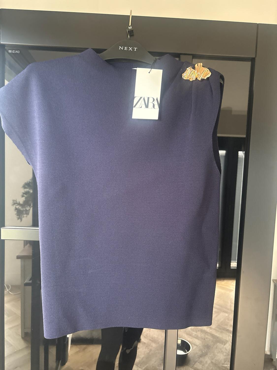 Navy blue sleeveless knitted top with gold-coloured applique, hanging on a hanger in front of glass. Text: EN12150, M, NEXT, ZARA.