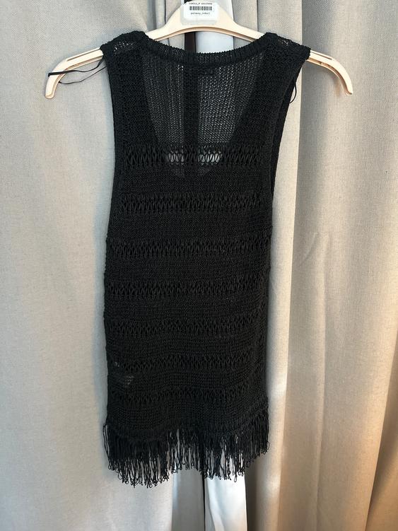 Black open-knit dress with fringe at the hem, hanging on a hanger against a backdrop of beige curtains.