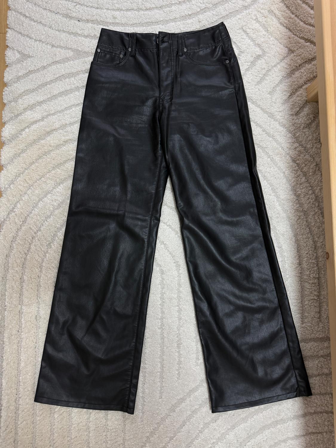 Black faux leather trousers on a beige carpet with a geometric pattern. Straight cut and five visible pockets.