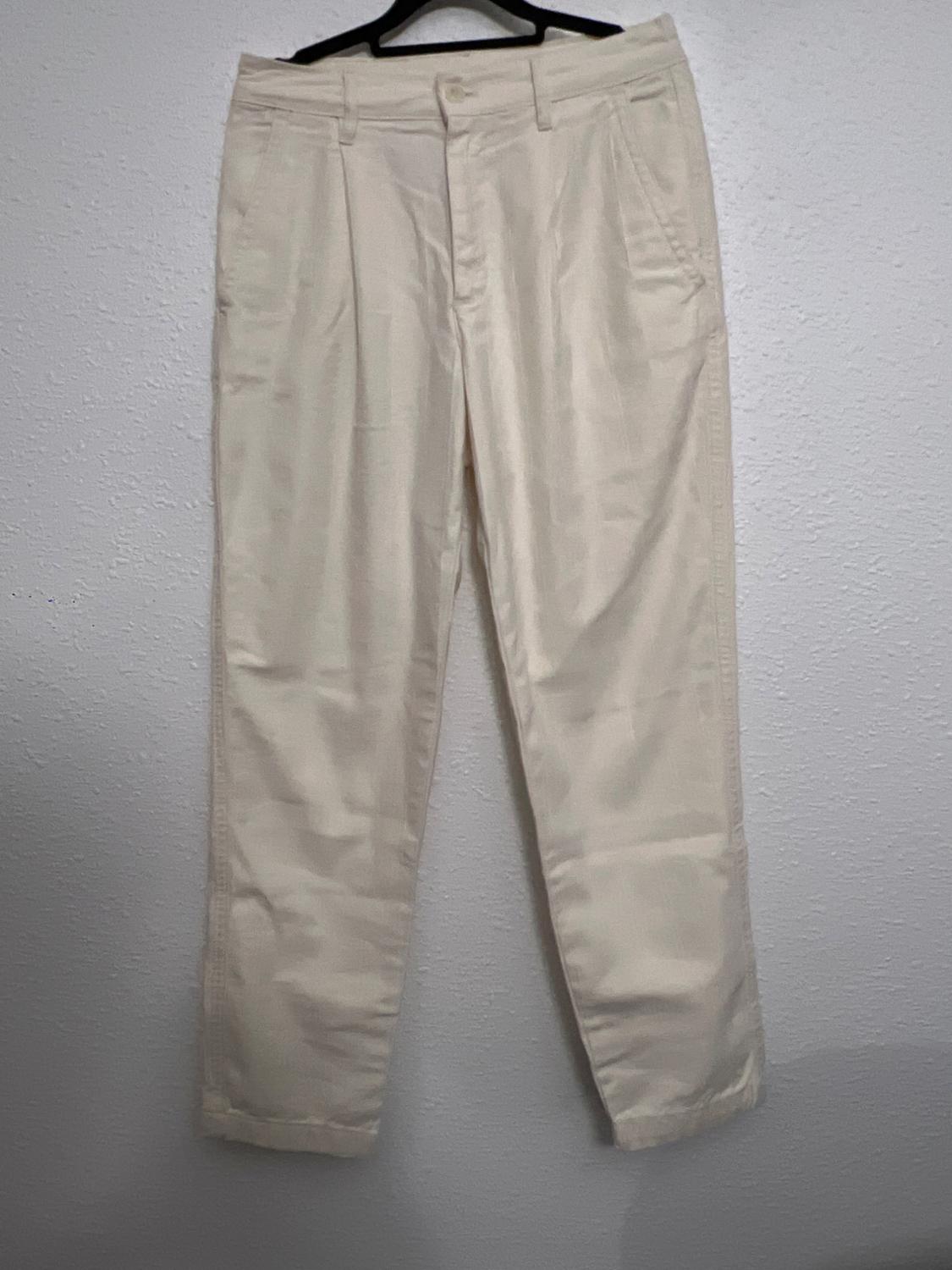 Beige trousers hanging on a black hanger against a white wall. Classic design with pleats and side pockets.