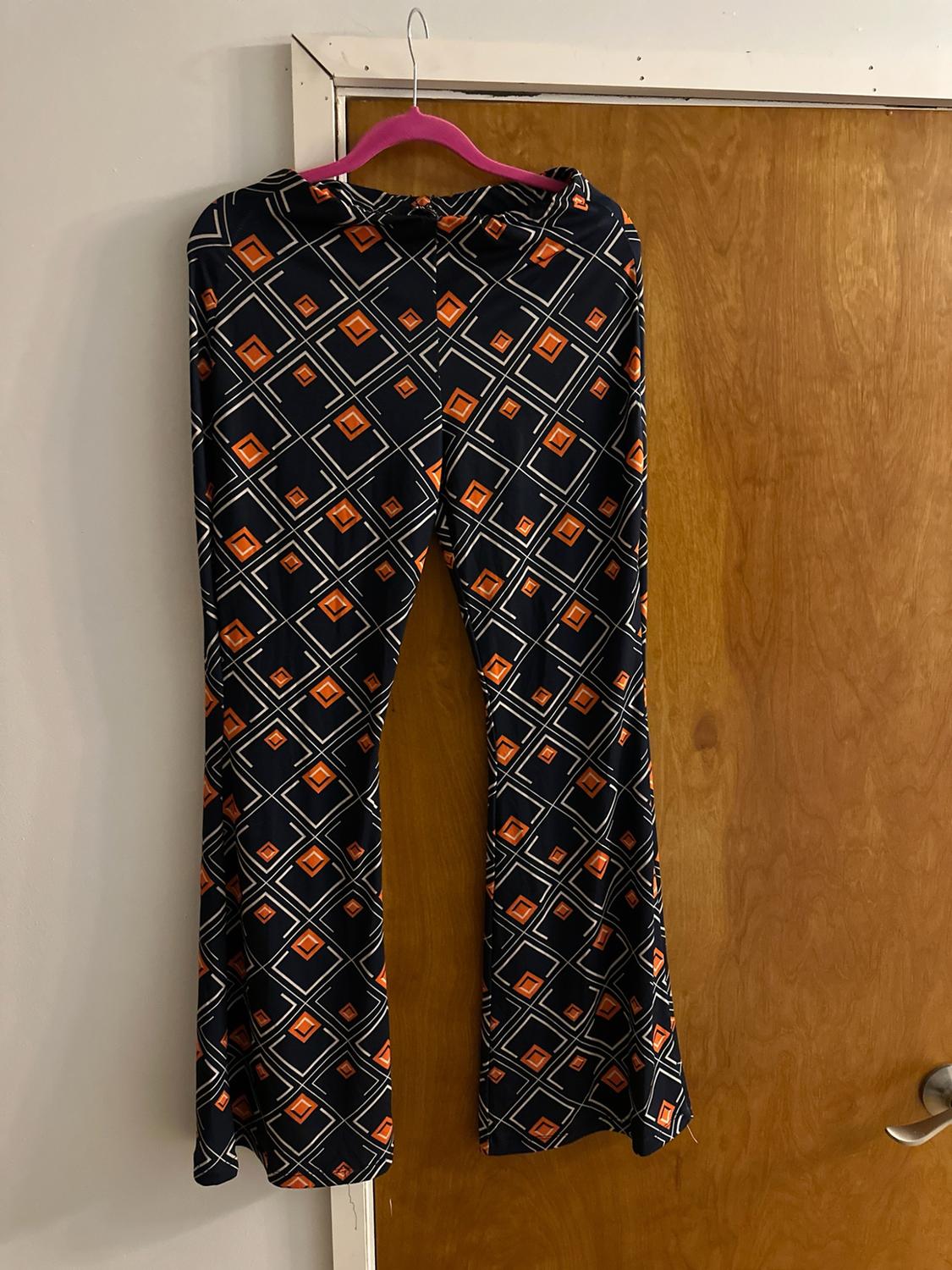 Flared trousers with a white and orange geometric square print on a dark background, hanging on a pink coat hanger on a door.