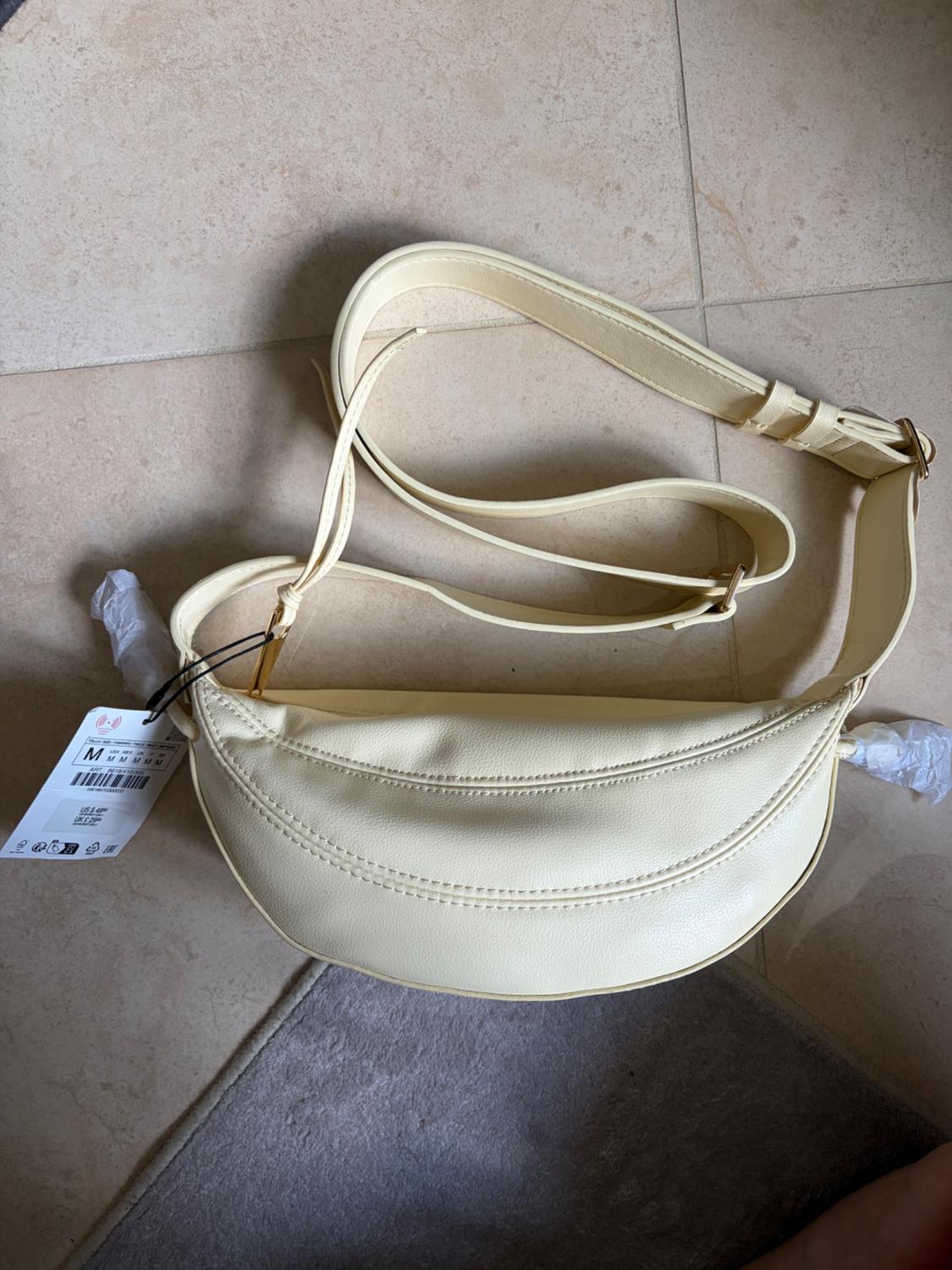 Light yellow crescent-shaped crossbody bag on a tiled floor. It has an adjustable strap and a label with the text M.