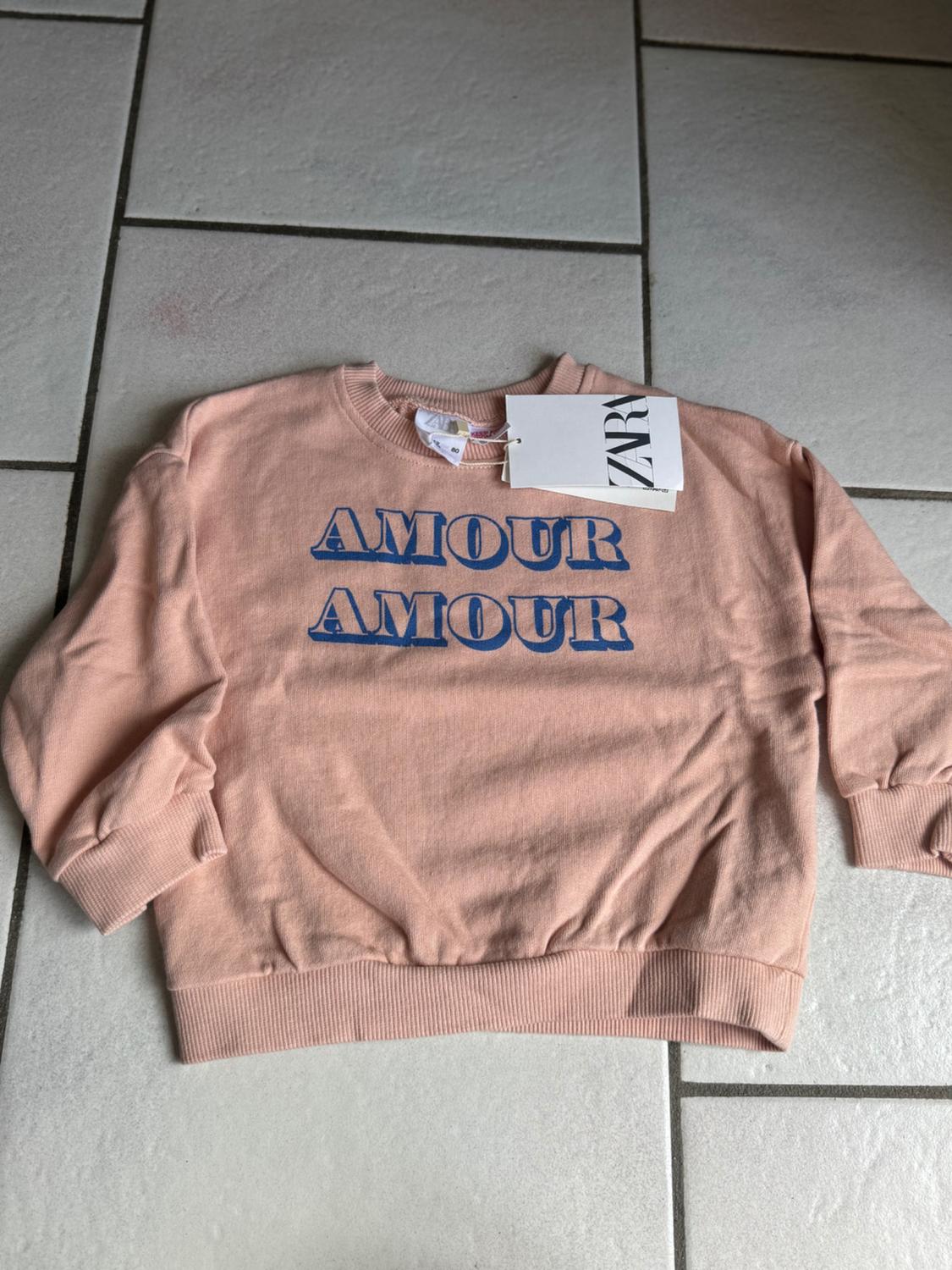 Pink long-sleeved sweatshirt on a tiled floor. Includes the blue text "AMOUR AMOUR" on the chest and a ZARA label on the neck.