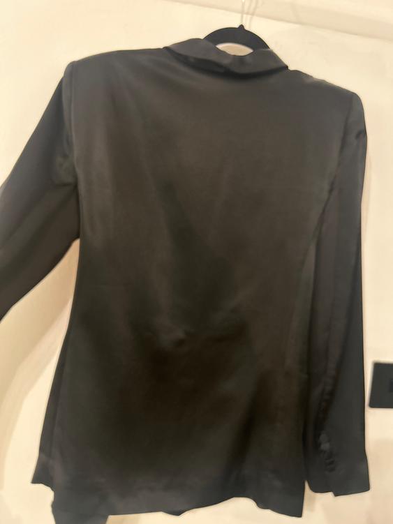 Black jacket hanging on a hanger, viewed from behind. The fabric appears soft and has a smooth finish. White background.