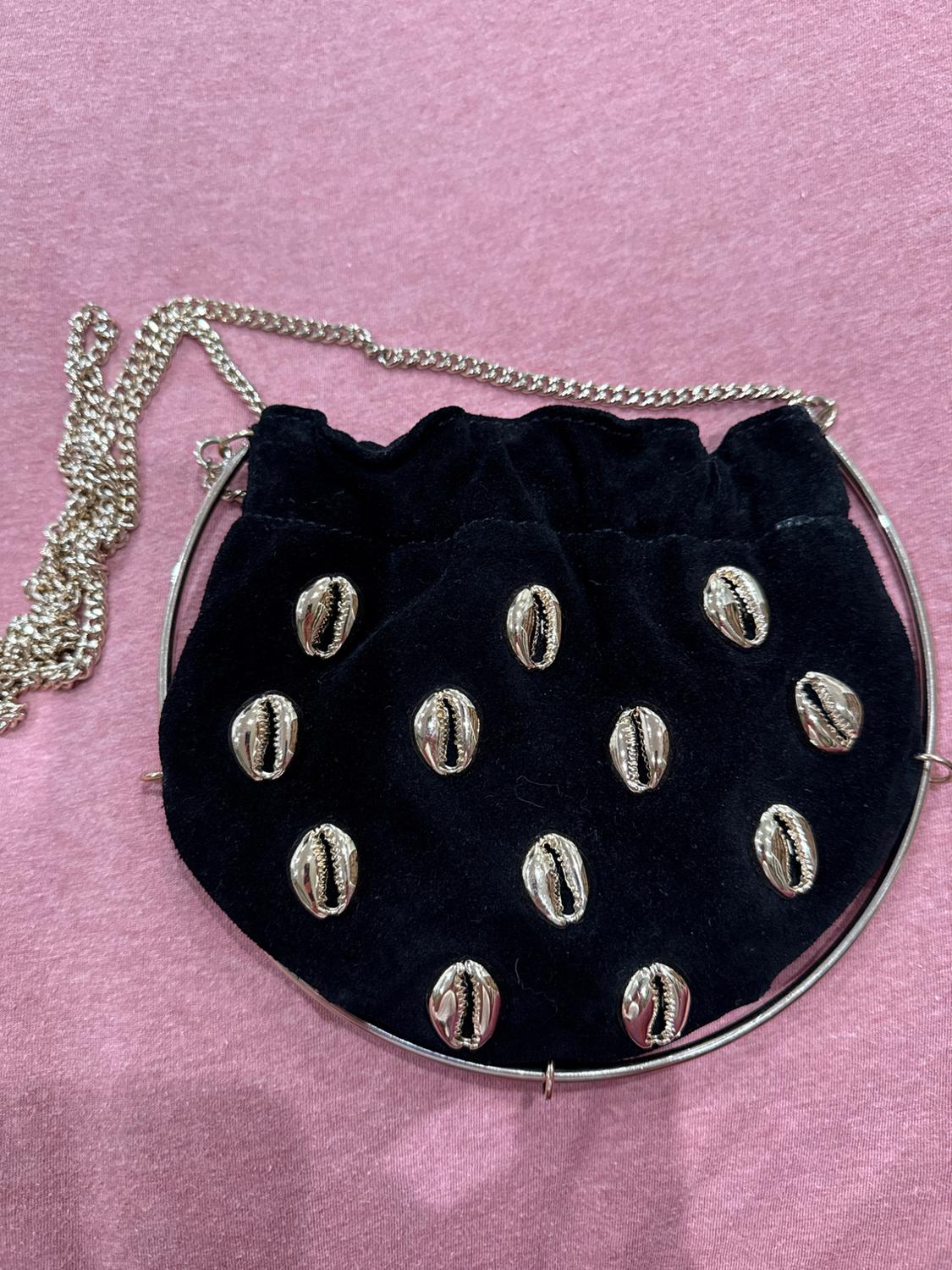 Black handbag with decorative metal shells and silver chain on a pink background.