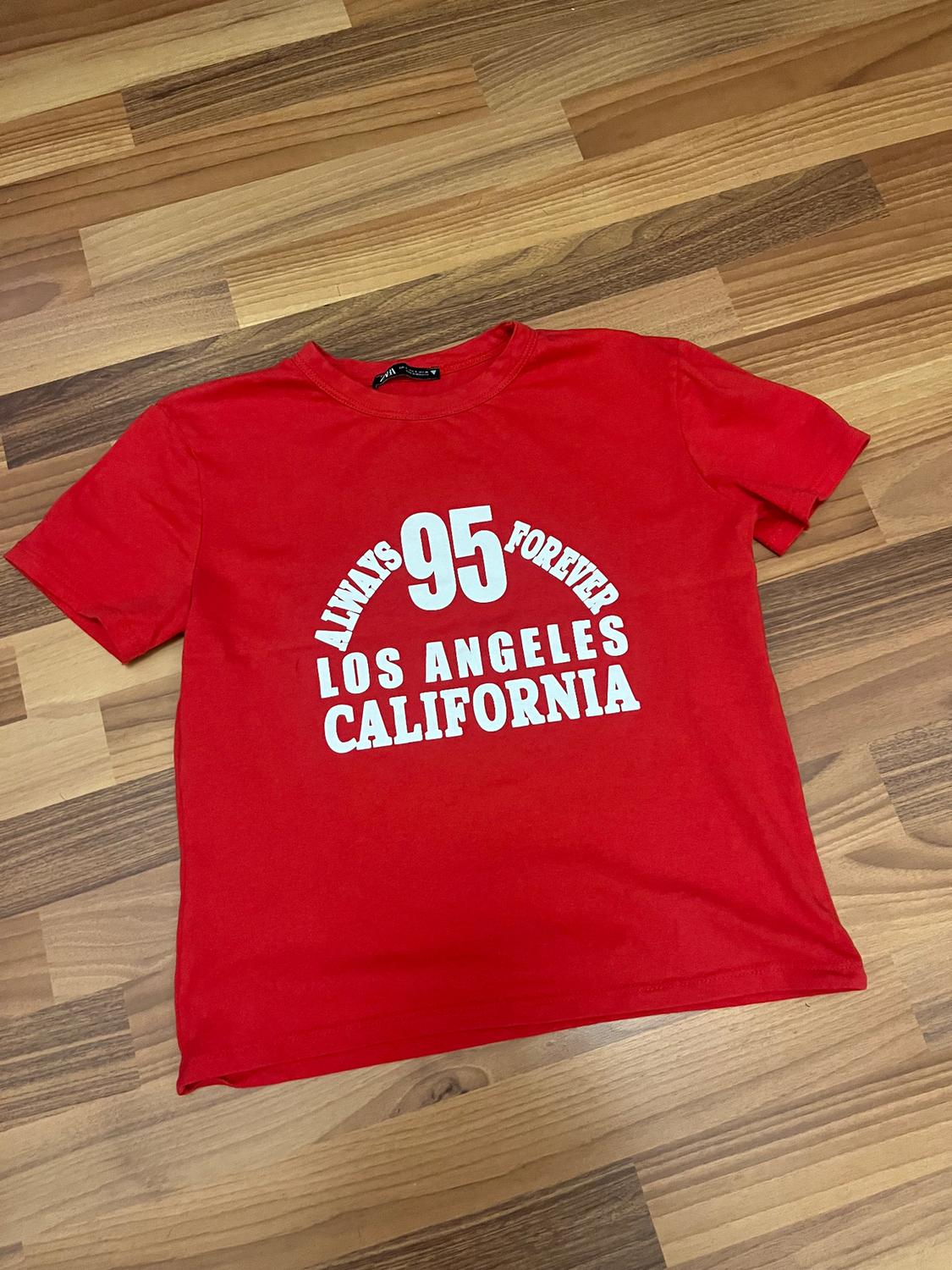 Red T-shirt with white text "ALWAYS 95 FOREVER LOS ANGELES CALIFORNIA" on a wooden floor.