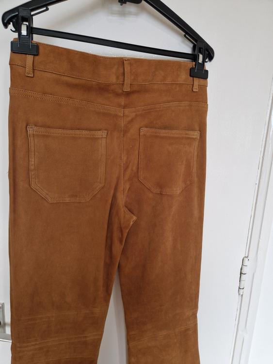 Brown suede trousers hanging on a black hanger, showing the back pockets and detailed stitching. White background.