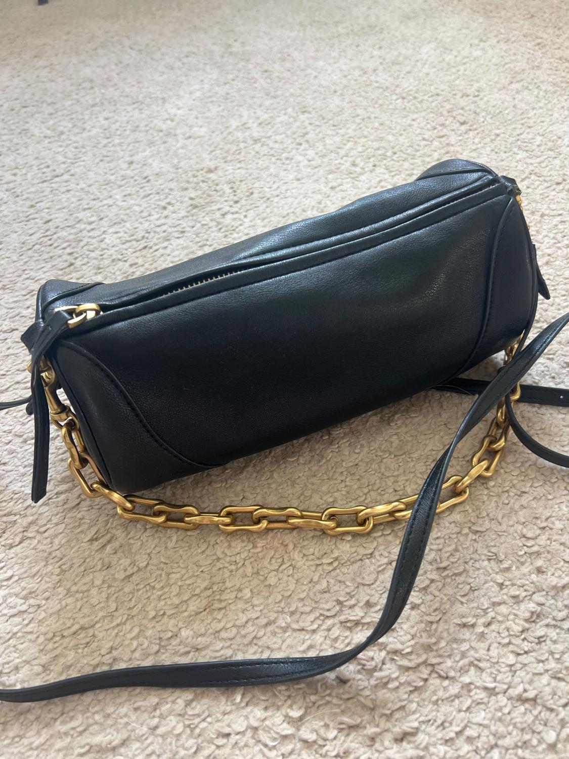 Black leather handbag with gold chain on beige carpet. Cylindrical design with zip fastening and adjustable strap.