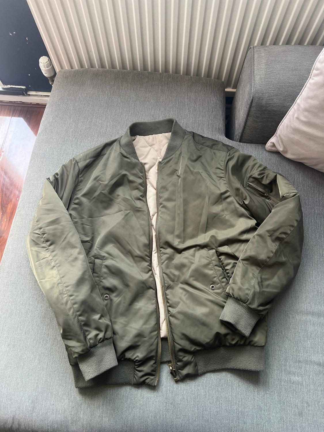 Olive green bomber jacket laid open on a grey sofa. Its pale quilted inner lining, ribbed cuffs and sleeve pocket stand out.
