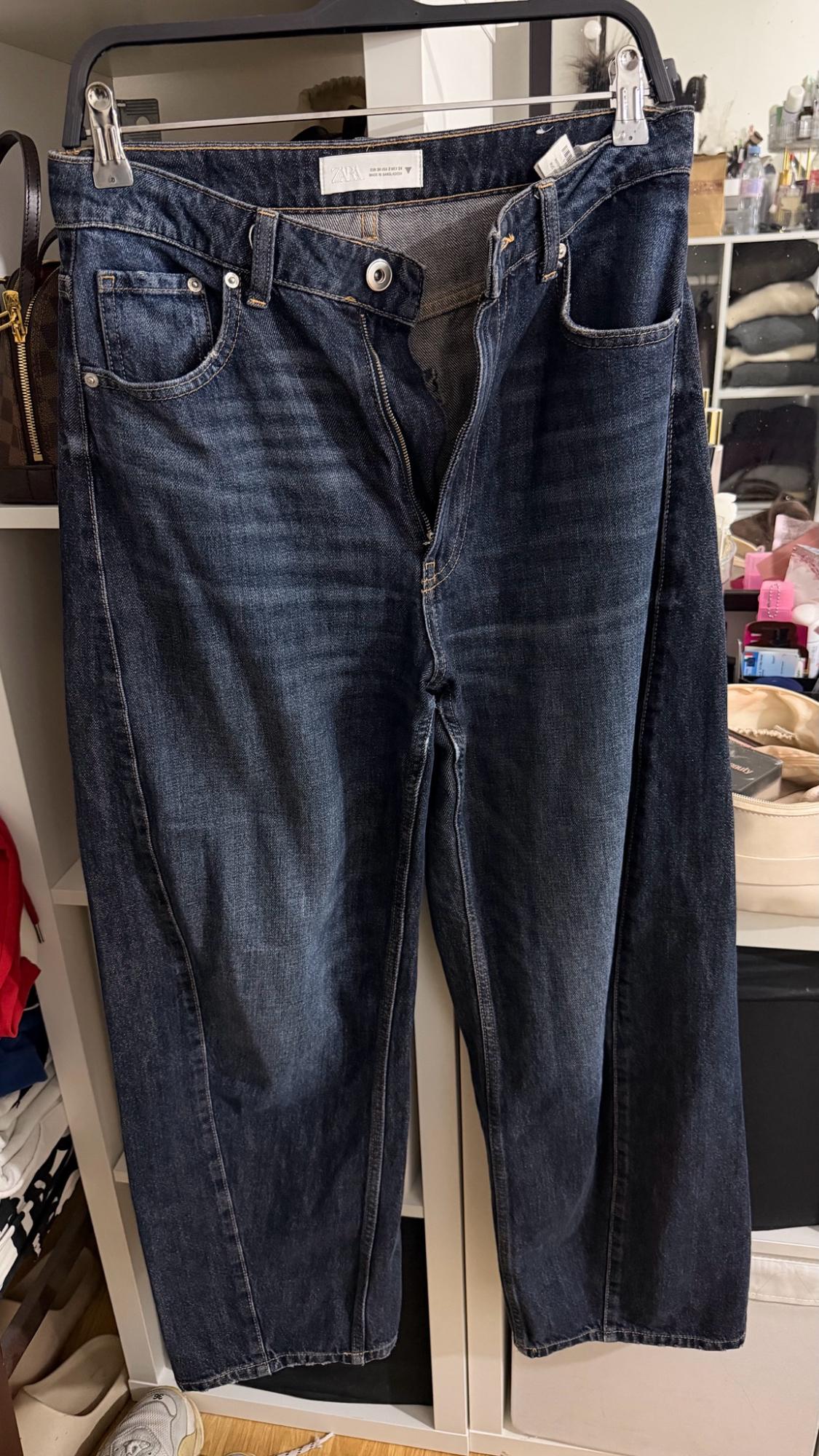 Dark blue jeans hanging on a hanger. Tag with the text ZARA. Casual setting in a wardrobe with shelves in the background.