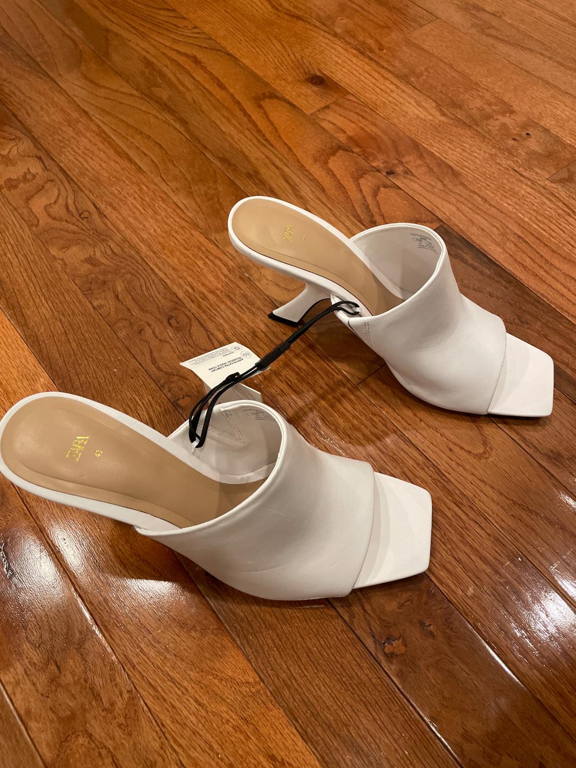 White high-heel shoes with a square toe on a wooden floor. A hangtag is visible on one of the shoes.
