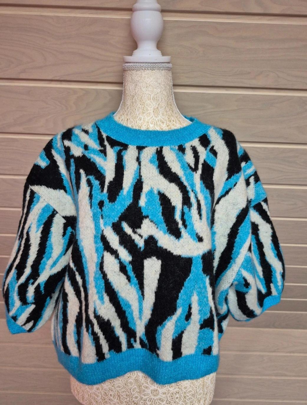 Mannequin wearing a short-sleeved knitted jumper with a zebra print in blue, white and black, in front of a light wooden wall.