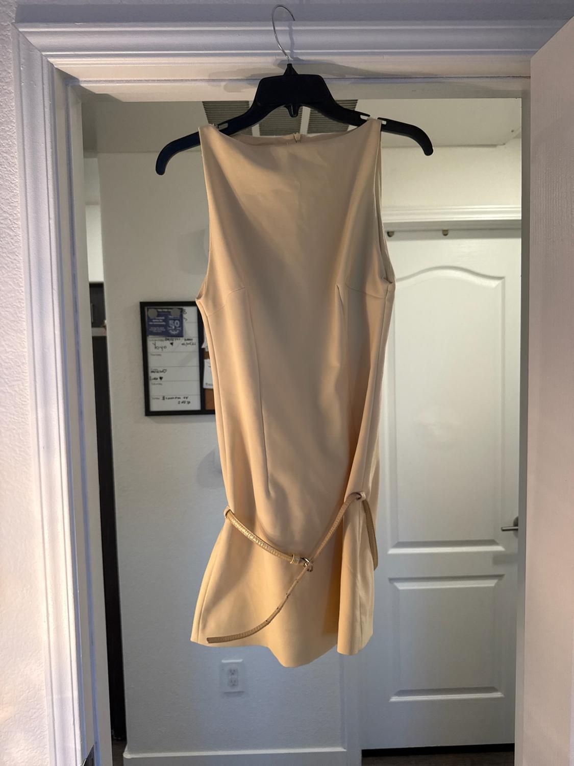 Short beige sleeveless dress with a thin belt, hanging on a black hanger from a white door frame in a home setting.