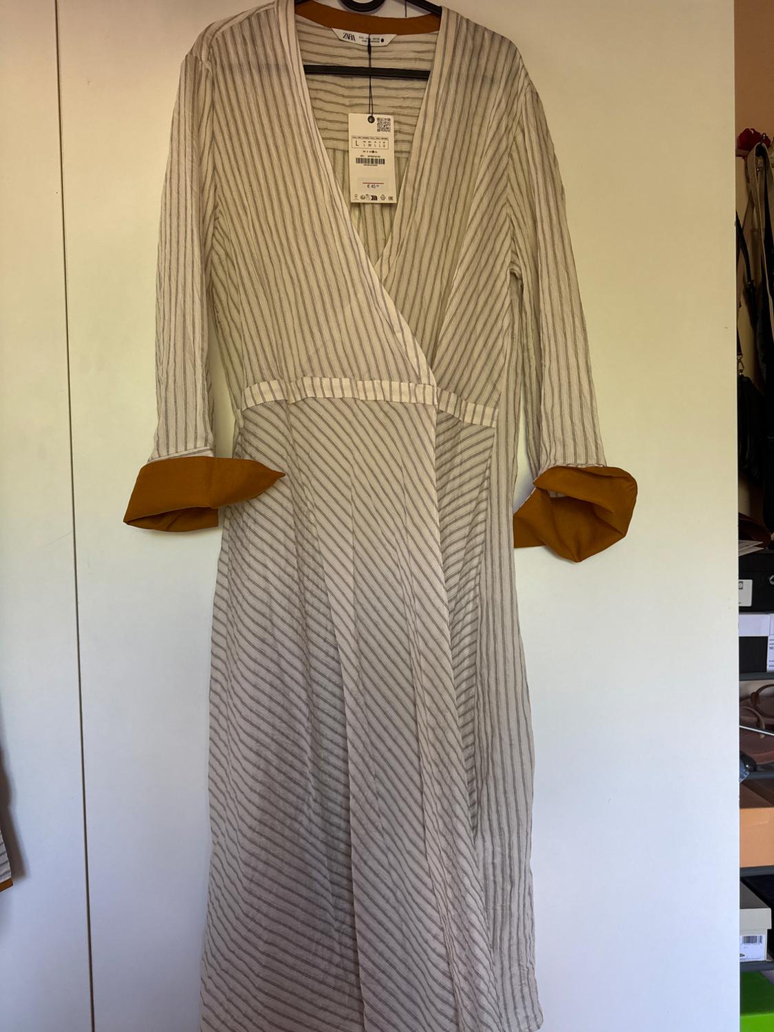 Long dress with vertical stripes in beige and white tones, with brown cuffs, hanging on a hanger against a white background. Label visible.