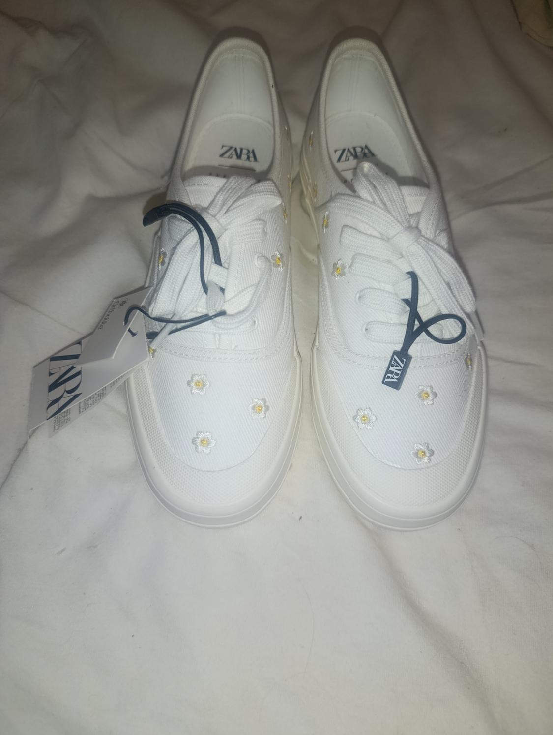 Pair of white trainers with small embroidered daisies on white fabric. ZARA is printed on the insoles and labels.
