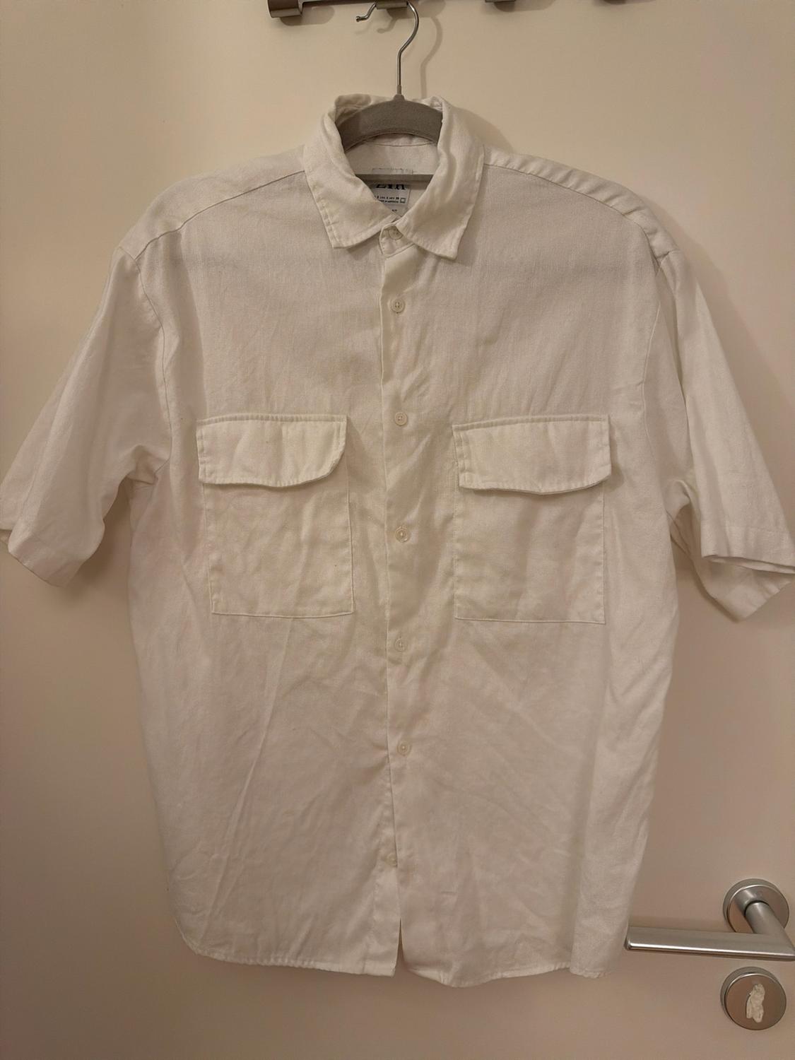 White short-sleeved button-down shirt with two front pockets, hanging on a hanger over a light-coloured door with a metal handle.