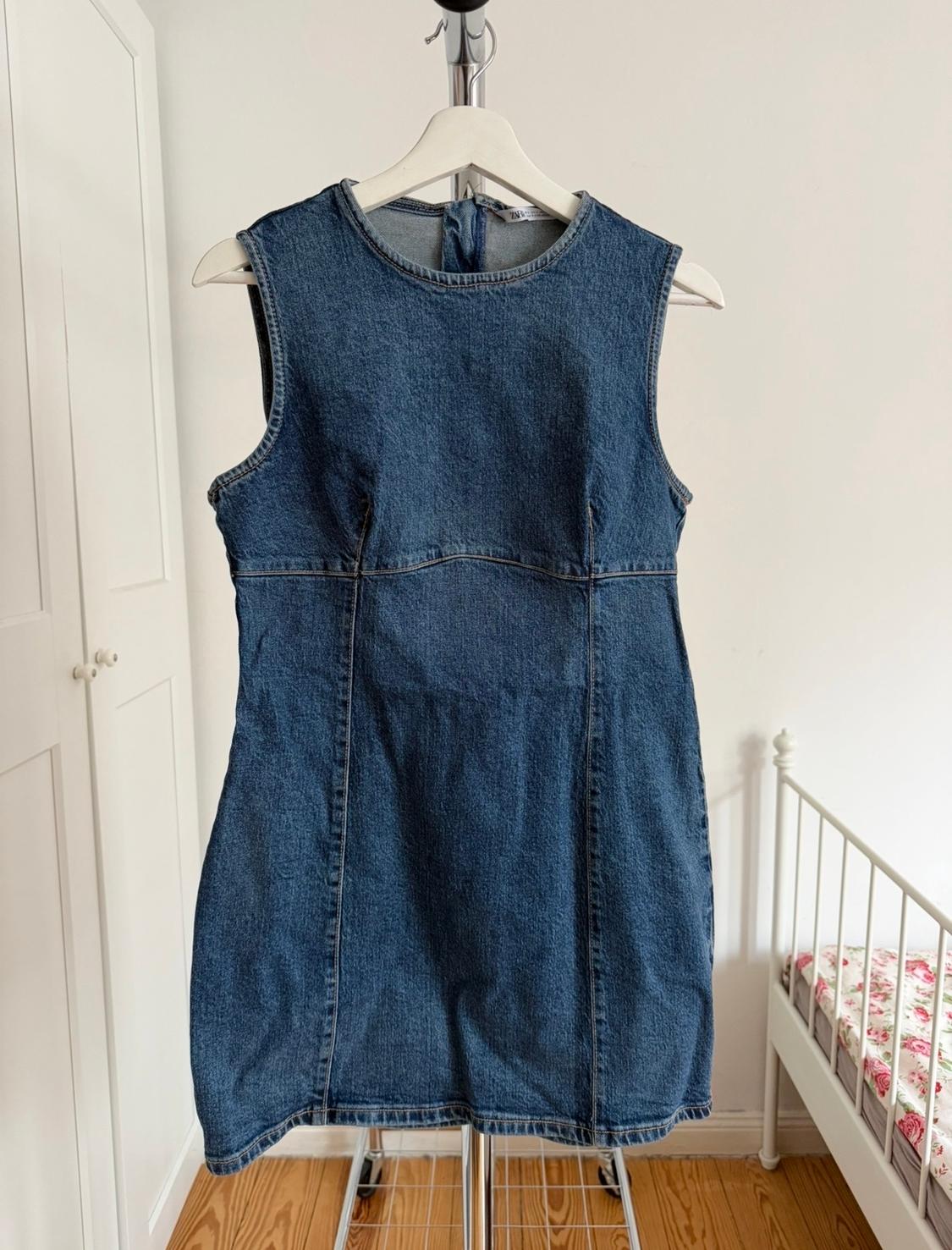 Αμάνικο φόρεμα από denim κρεμασμένο σε μια λευκή κρεμάστρα σε ένα δωμάτιο με μεταλλικό κρεβάτι και ξύλινο πάτωμα.