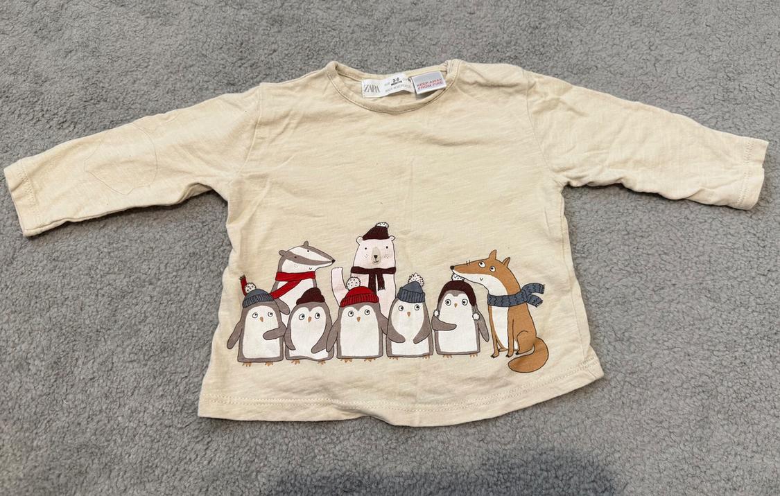Beige long-sleeved T-shirt with illustrations of penguins, a polar bear, and a fox, all wearing scarves and winter hats.