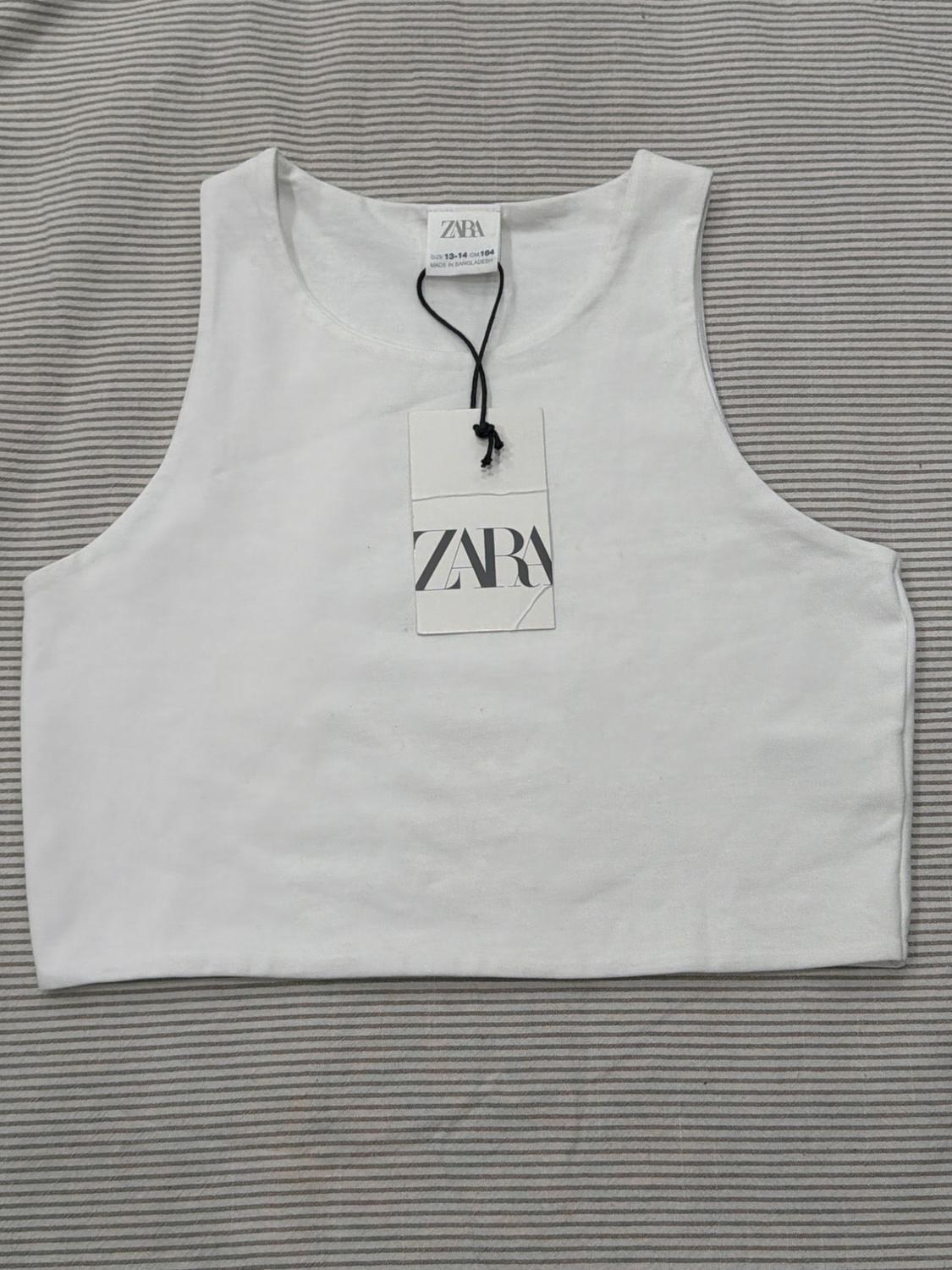 White sleeveless top over a gray striped background. Hang tag with the text "ZARA" visible in the center of the top.