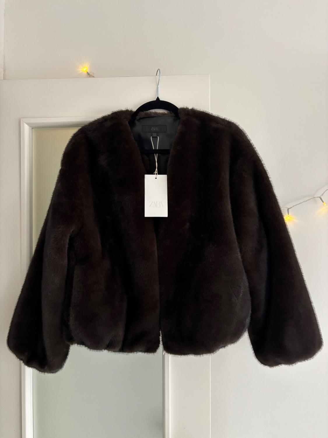 Dark brown faux fur coat hanging on a black hanger with a white Zara label, white background with yellow lights.