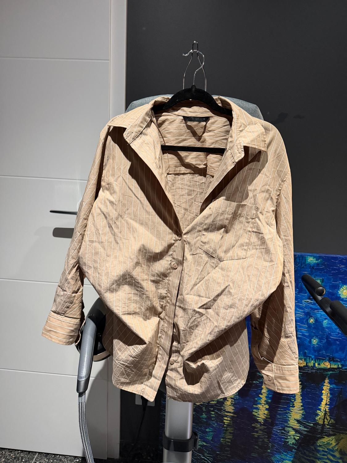 Beige pinstripe shirt hanging on a hanger over a grey chair, with a black wall background and a colourful painting.
