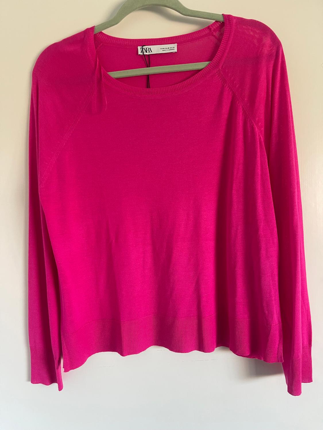 Fuchsia pink long-sleeved jumper hanging on a green hanger. Visible label on the collar. White background.