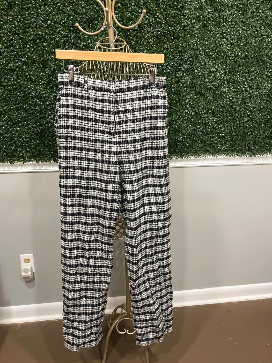 Black and white plaid pants hanging on a golden rack in front of a green leaf wall.