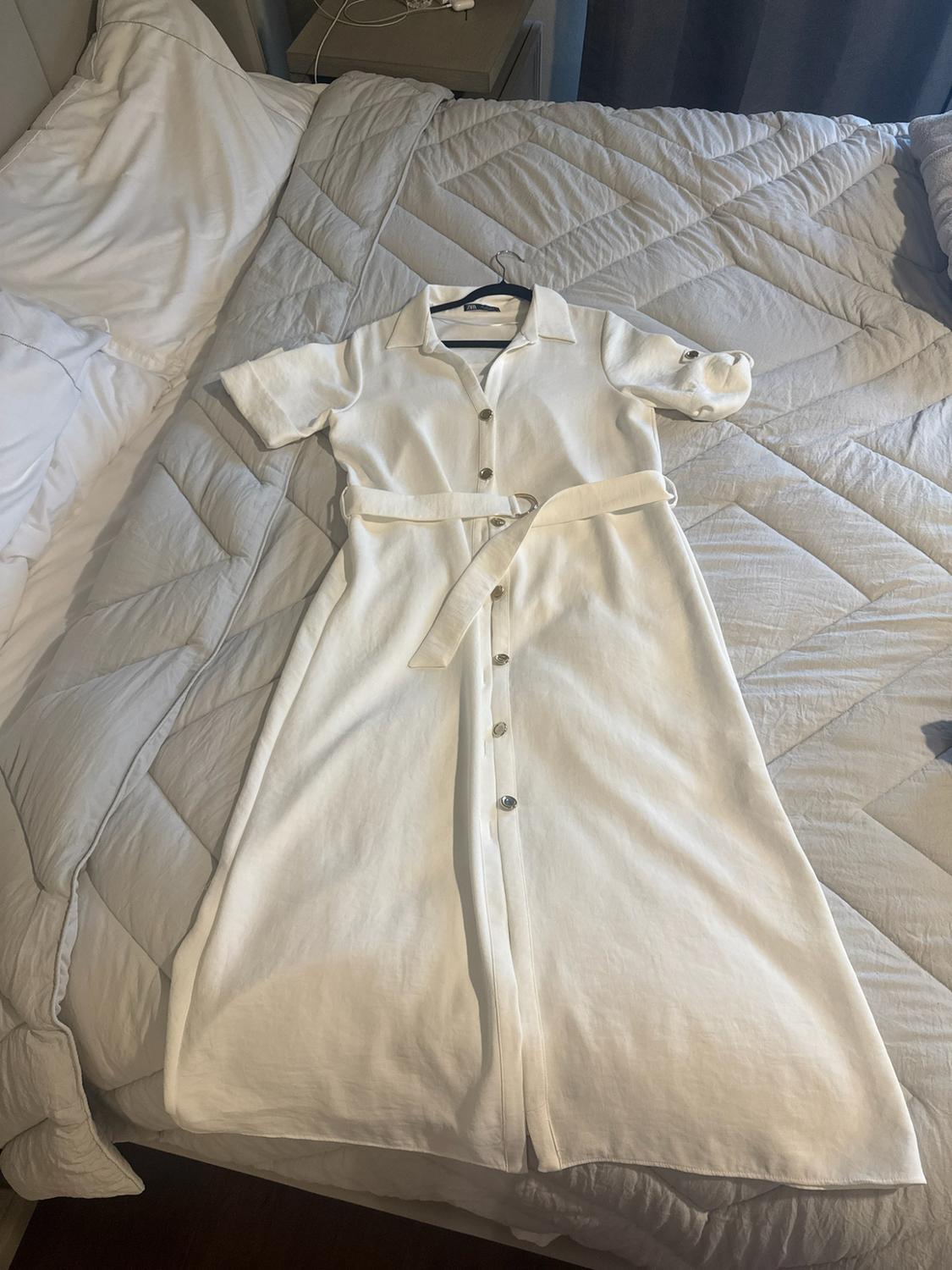 White short-sleeved shirt dress with buttons and a belt, laid out on a bed with a light-coloured duvet. Casual style in a relaxed setting.