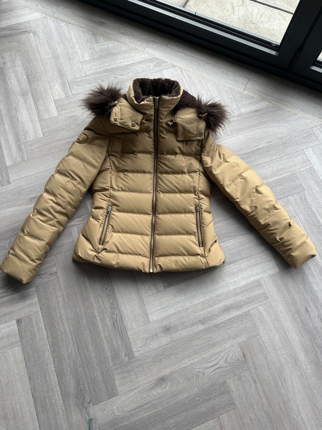 Beige padded jacket with hood and dark faux fur trim, on light grey wooden floor.