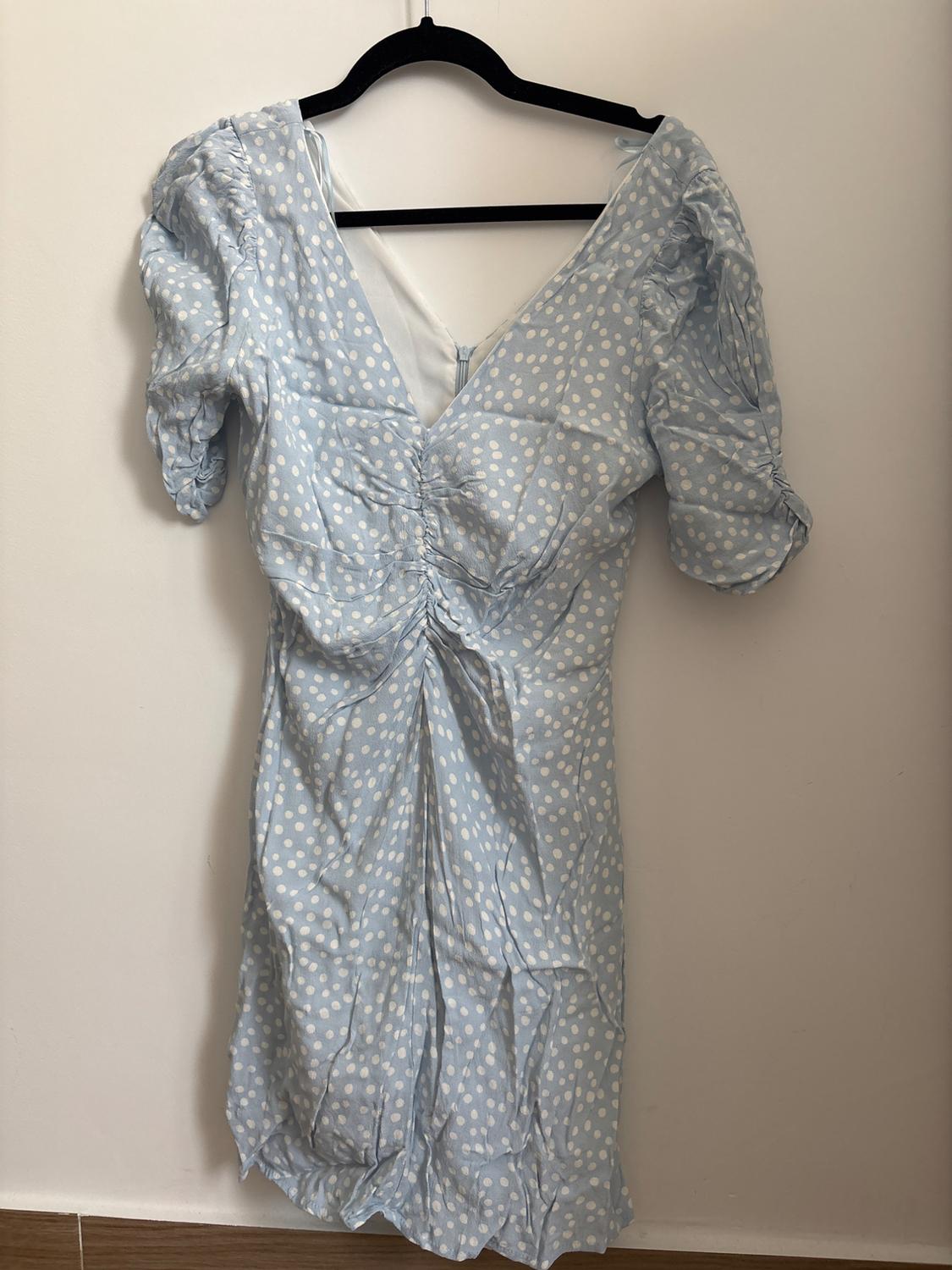 Light blue dress with white polka dots on a black hanger against a white wall. It features a V-neck, front ruching and short puff sleeves.