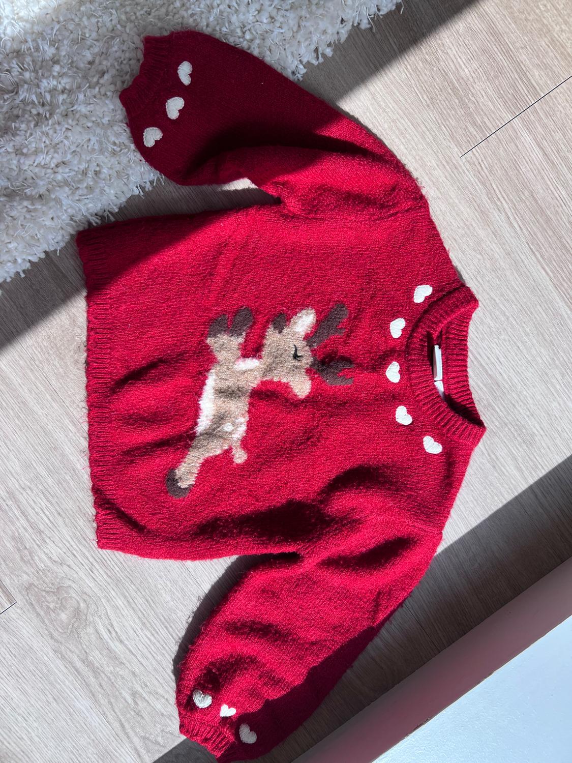 Red knit sweater with a reindeer motif in the center and white hearts around the neckline and on the sleeves, on a wooden floor.