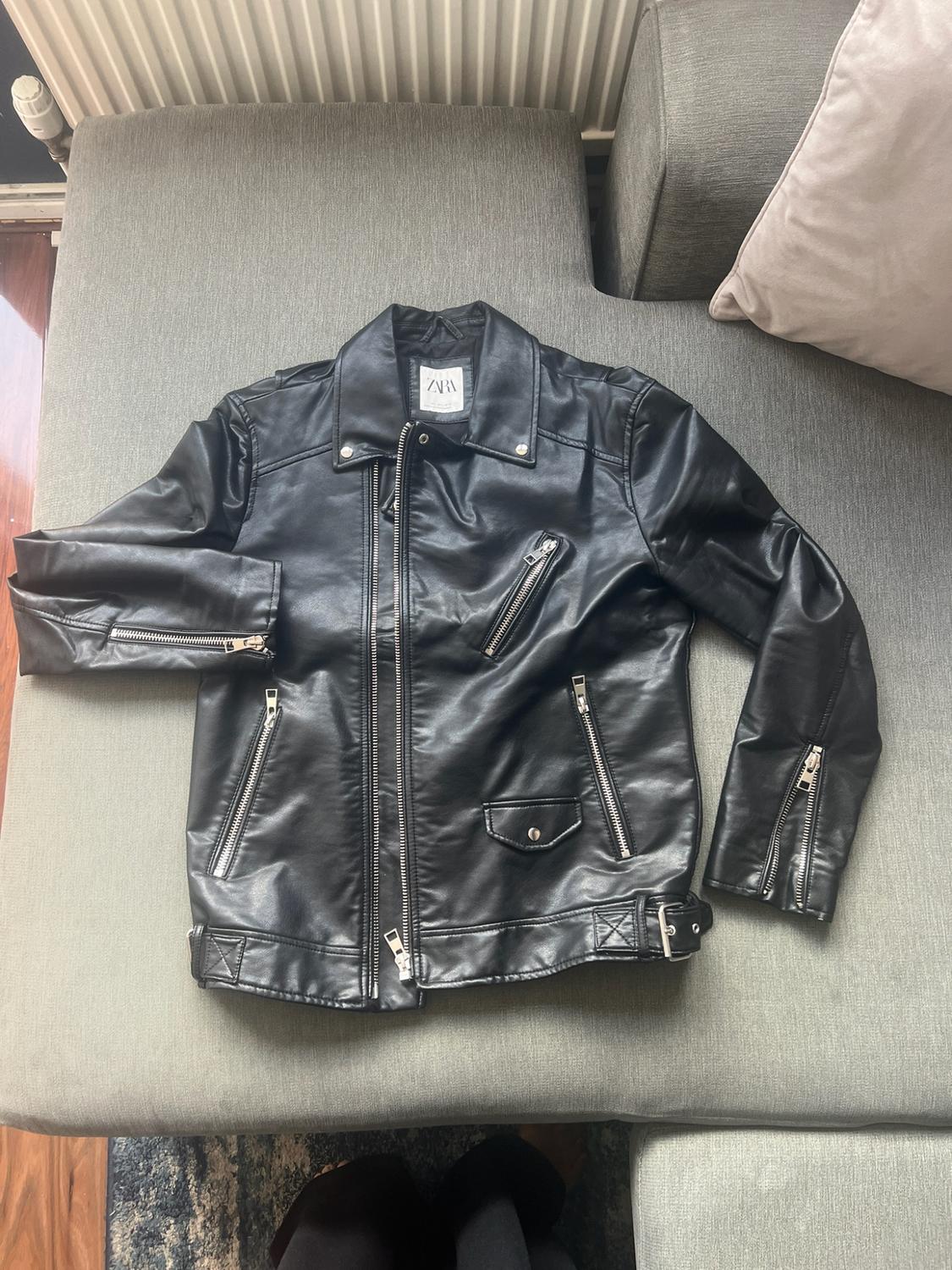 Black biker jacket with silver zips on a grey sofa in a relaxed setting. Label with the text ZARA.