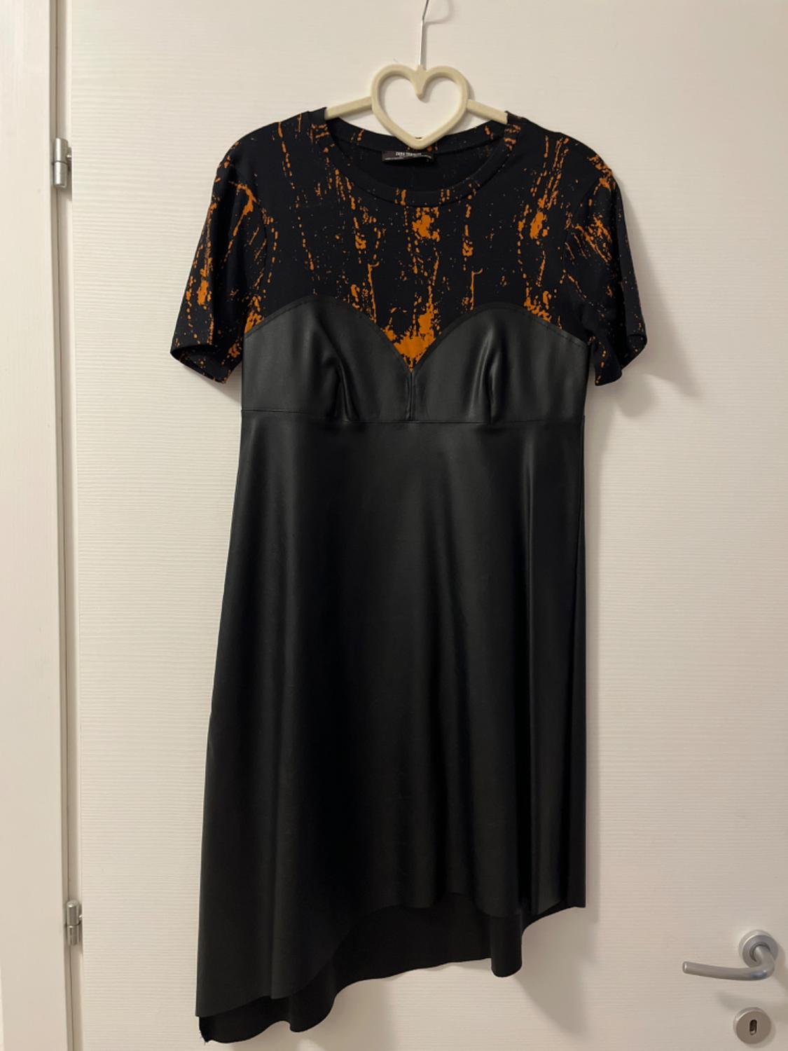 Short-sleeved dress in white doorway. Orange patterned top and glossy black skirt with an asymmetrical hem.