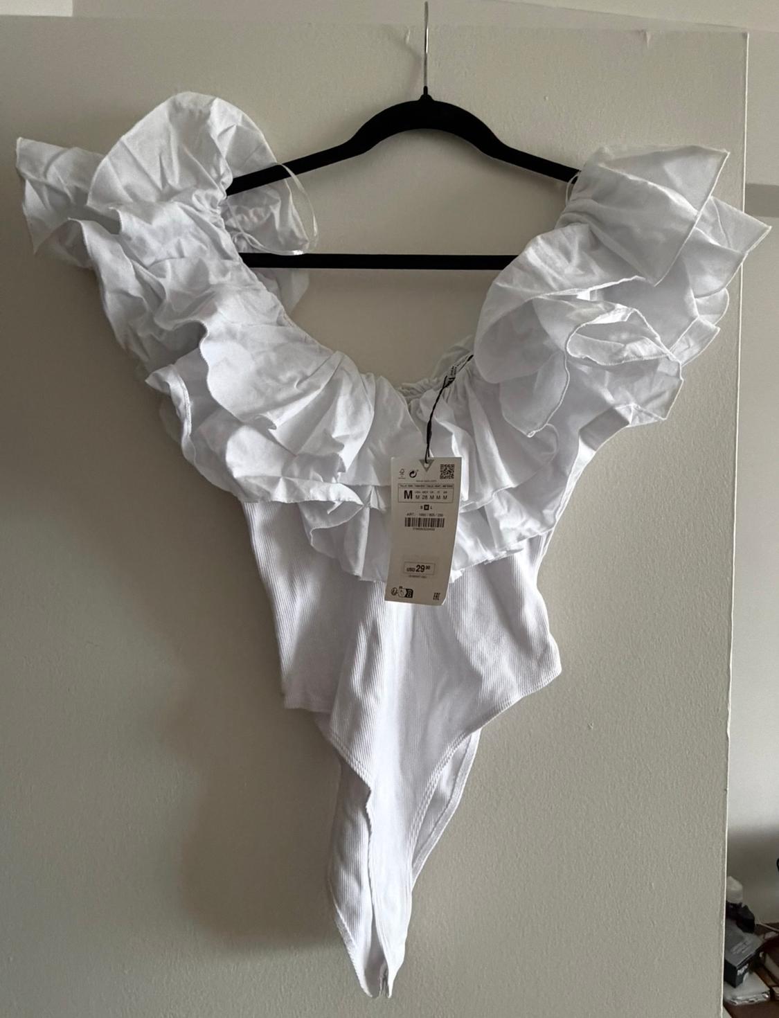 White bodysuit with ruffled shoulders, hanging on a black hanger. Visible tag with product details.