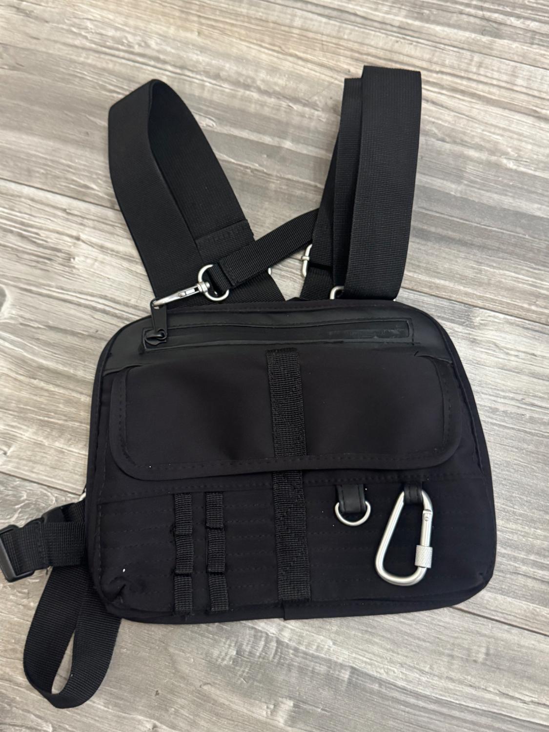 Black utility-style harness bag on a wooden floor. It has thick straps, a flap pocket, a zip and a silver carabiner.