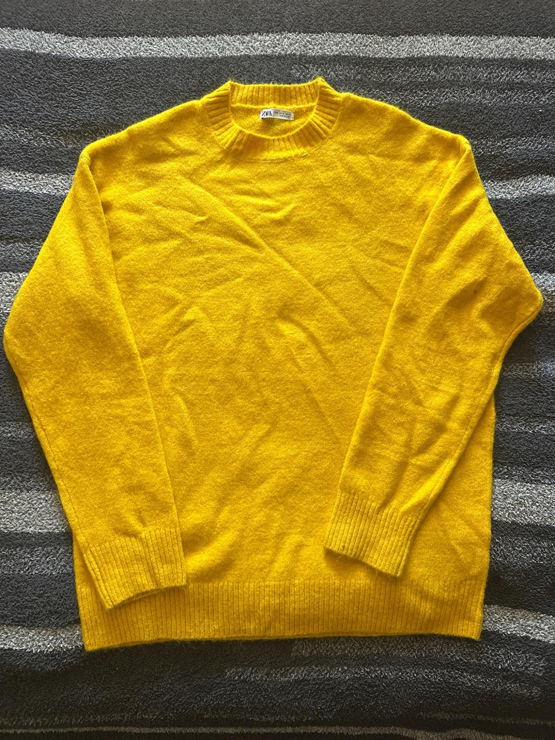Yellow knit sweater with long sleeves and a round neckline, on a surface of gray and white stripes. Label with the text ZARA.