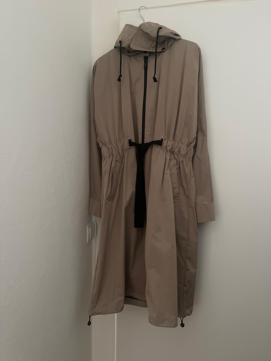 Beige coat with a hood and black drawstrings hanging on a white door. Long design with a cinched waist.