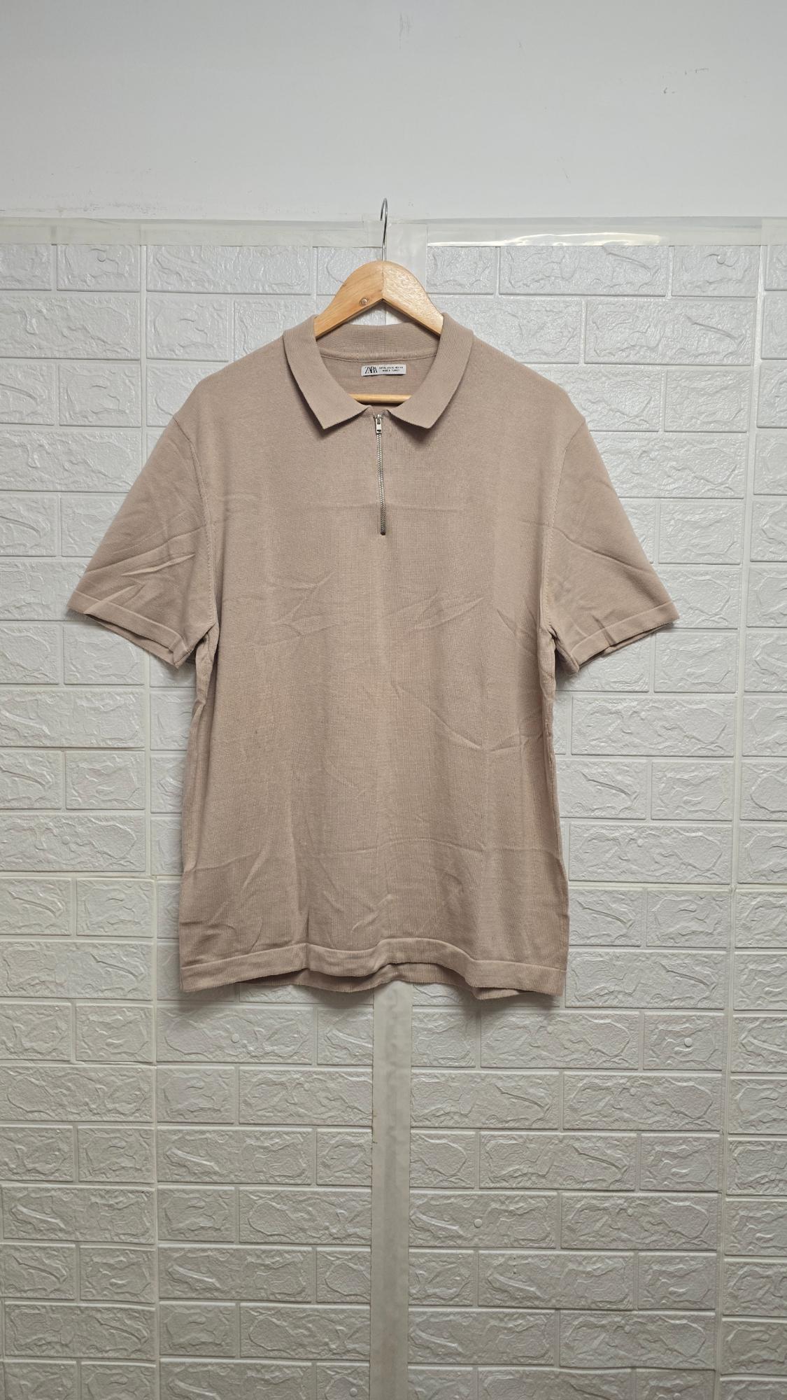 Beige polo shirt with a zip hanging on a wooden coat hanger, against a white brick-textured wall background.