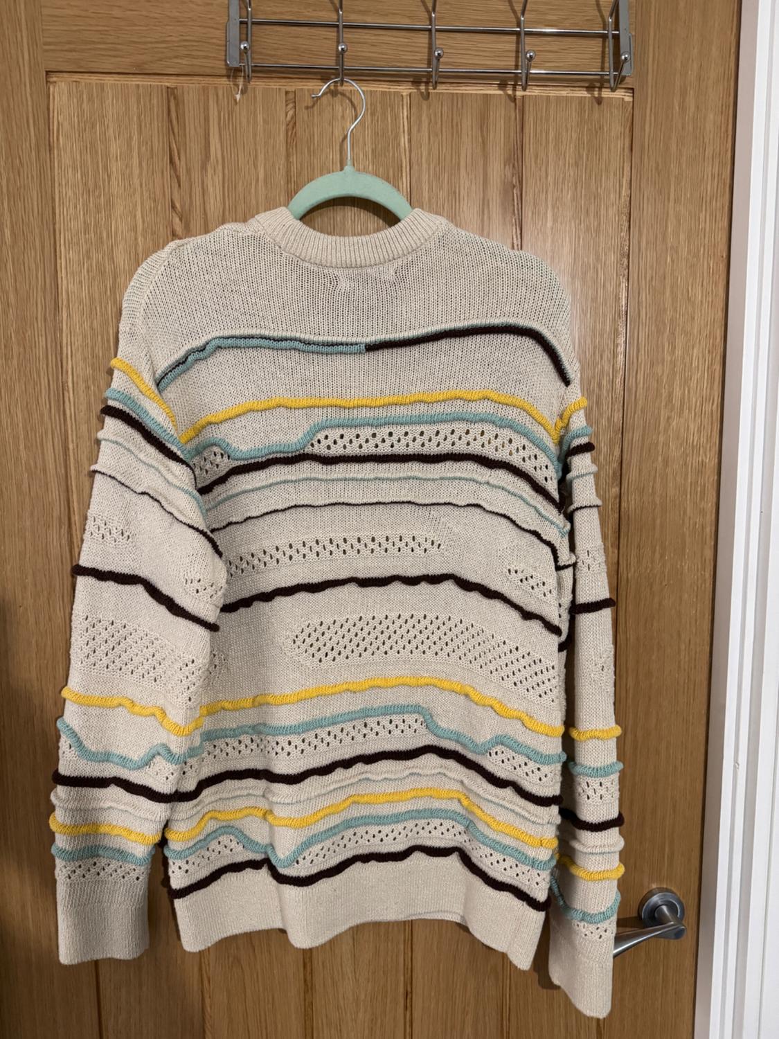 Beige knitted jumper with wavy stripes in brown, yellow, and light blue, hanging on a wooden door.
