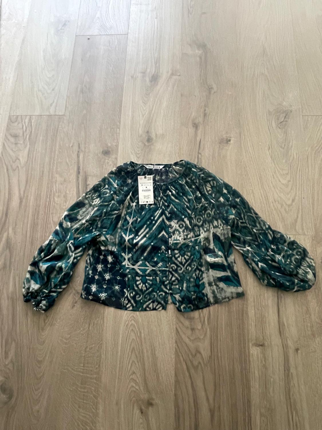 Long-sleeved blouse with geometric print in shades of green and blue on a light wood floor. Visible label on the neck.
