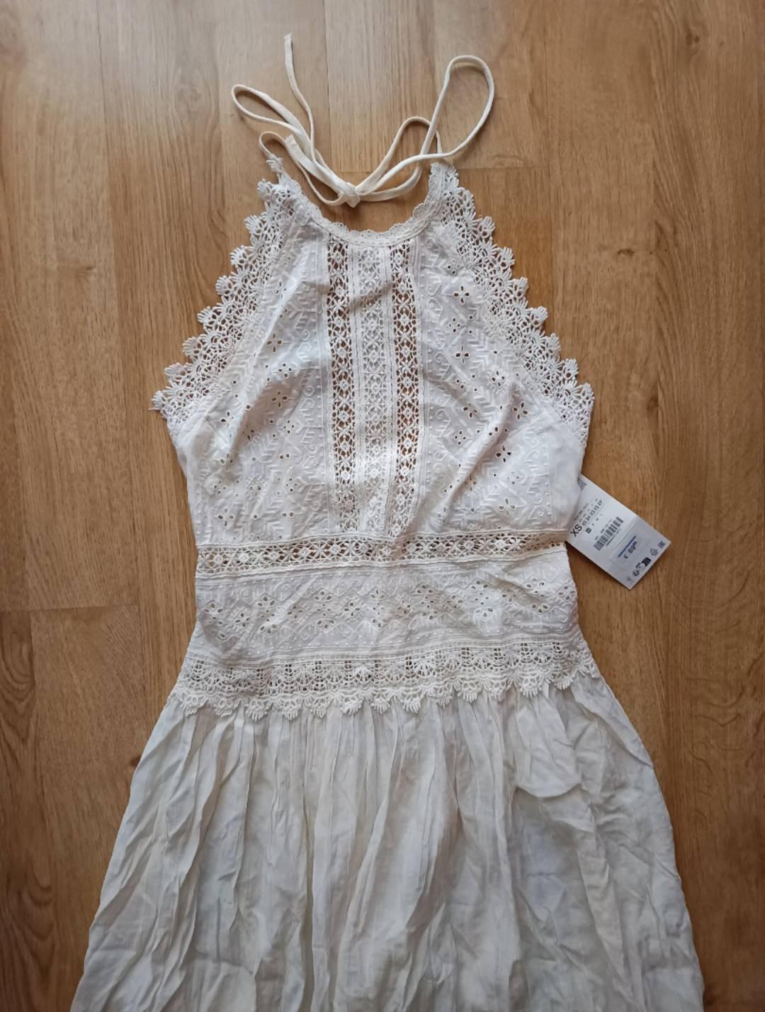 Relaxed-fit white halter-neck dress with embroidery and lace, on a wooden floor. An XS label is visible.