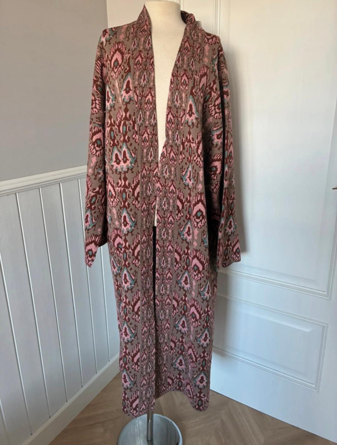 Mannequin in a long geometric print dress in pink and brown tones, in a room with white walls and a light wooden floor.