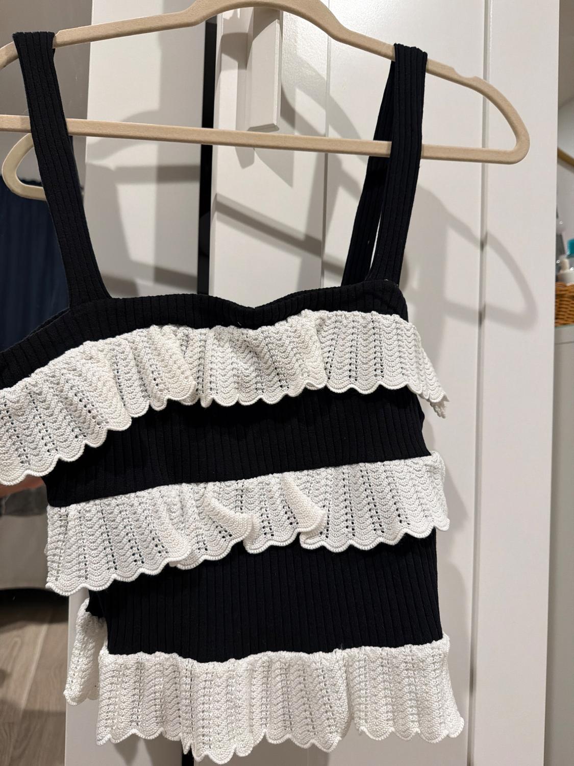 Black strappy top with layered white ruffles, hanging on a wooden hanger in front of a white wardrobe.