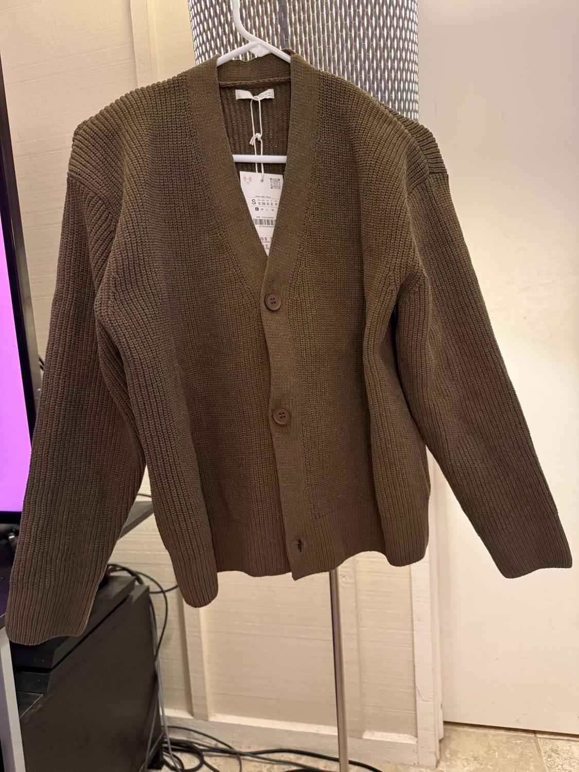 Olive-green knitted cardigan with front buttons hanging on a white hanger in a room. It has a visible label marked S.