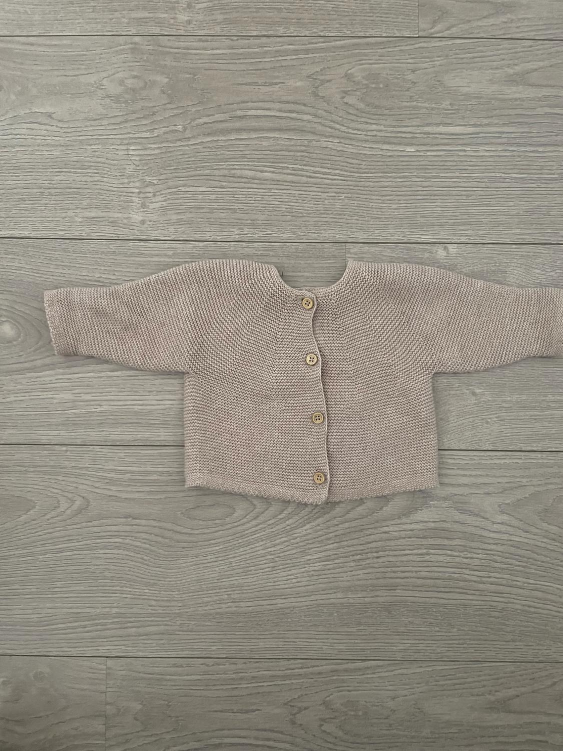 Beige knitted baby cardigan with wooden buttons, on a grey wooden floor with visible texture.