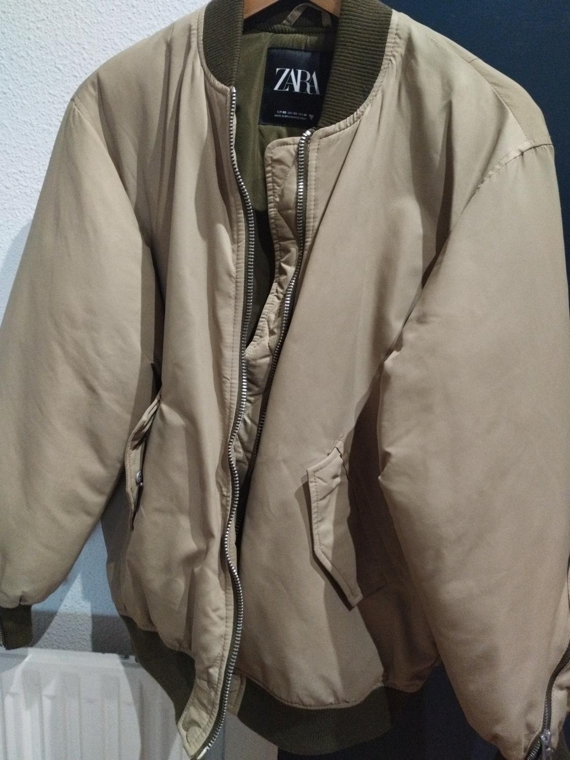 Beige bomber jacket hanging on a hanger, with a front zip and green elasticated cuffs. Visible label with text "ZARA".