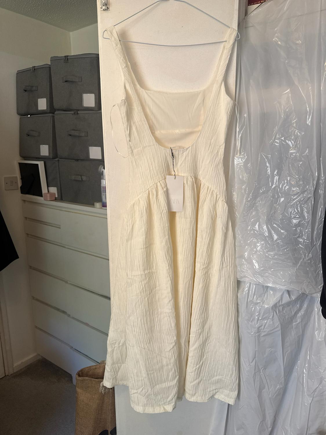 White textured strappy backless dress hanging in a room. Label with ZARA text.