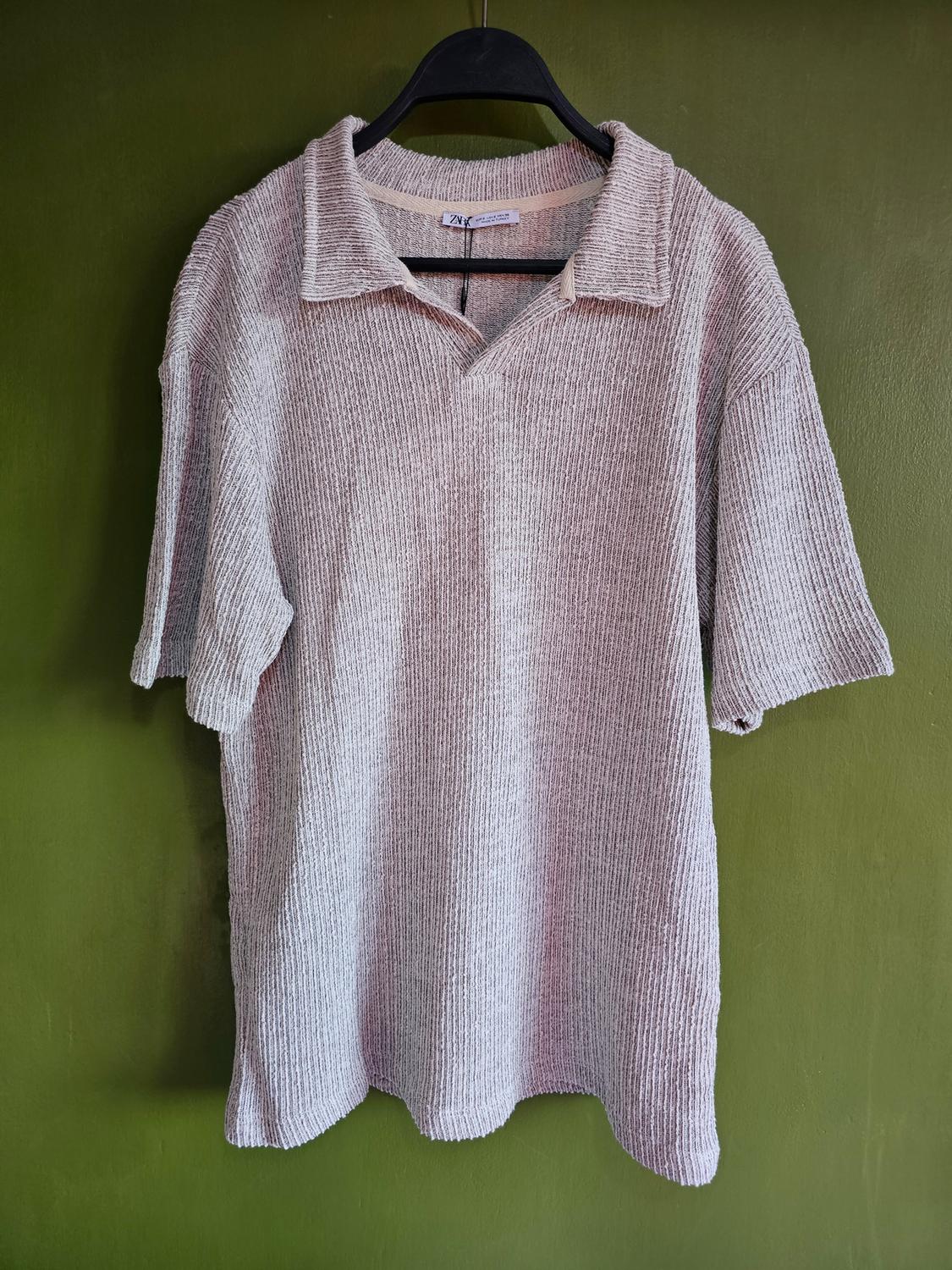 Beige knitted polo shirt hanging on a black hanger against a green background. Visible label with text "Zara".